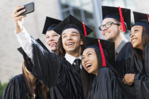 Multiracial group of six friends taking a selfie at graduation, in their black caps and gowns